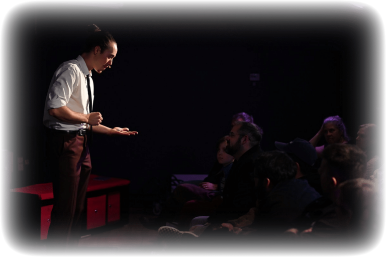 Boston stage magician and mentalist Eaden Marti holding out his hand with coins on it in front of a seated audience.